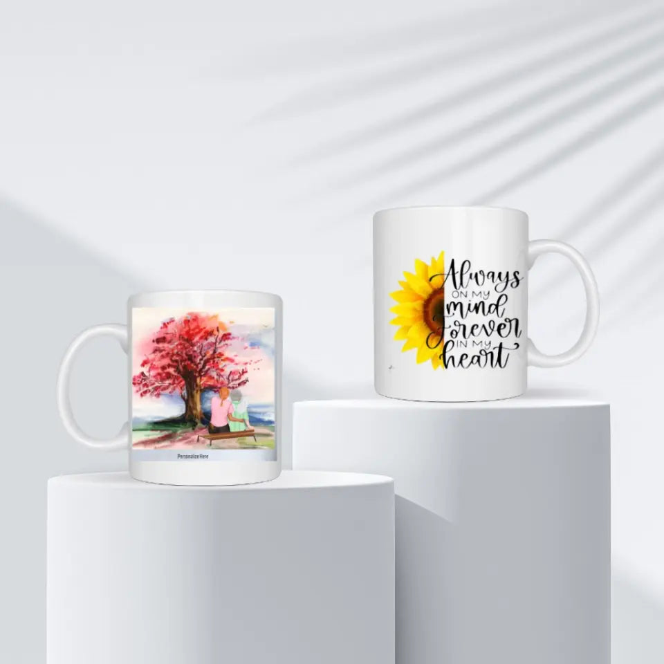 Two artistic coffee mugs on display, one with a serene tree painting and personalized text, the other with a vibrant sunflower and 'Always on my mind, forever in my heart' quote, available in 11oz & 15oz at Grief Relief Merch on www.griefreliefsupport.com.